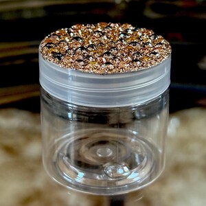 May include: A clear, cylindrical jar with a transparent lid. The lid is adorned with a sparkling, textured surface of gold and bronze-colored embellishments. The jar is empty, showcasing its clear plastic construction.