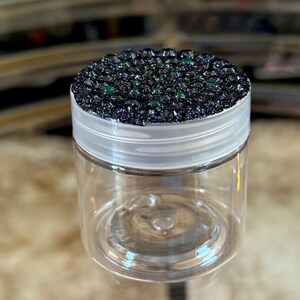 May include: A clear, empty jar with a white lid. The lid is covered in dark, sparkly material with small green accents. The jar is sitting on a reflective surface.