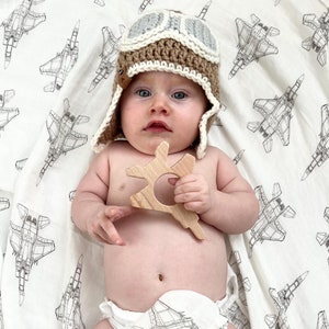 May include: A baby wearing a brown and gray crocheted aviator hat and a white diaper, holding a wooden teether shaped like a star. The baby is lying on a white blanket with a black and white pattern of fighter jets.
