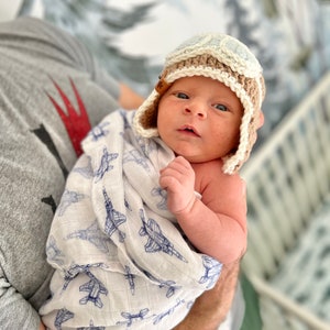 May include: A newborn baby wearing a crocheted aviator hat with blue and brown accents. The baby is wrapped in a white swaddle blanket with a blue airplane pattern.