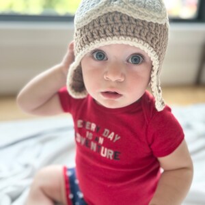 May include: A baby wearing a brown and white crocheted aviator hat with ear flaps, a red shirt with the text "Every Day is an Adventure", and blue and white star patterned shorts.