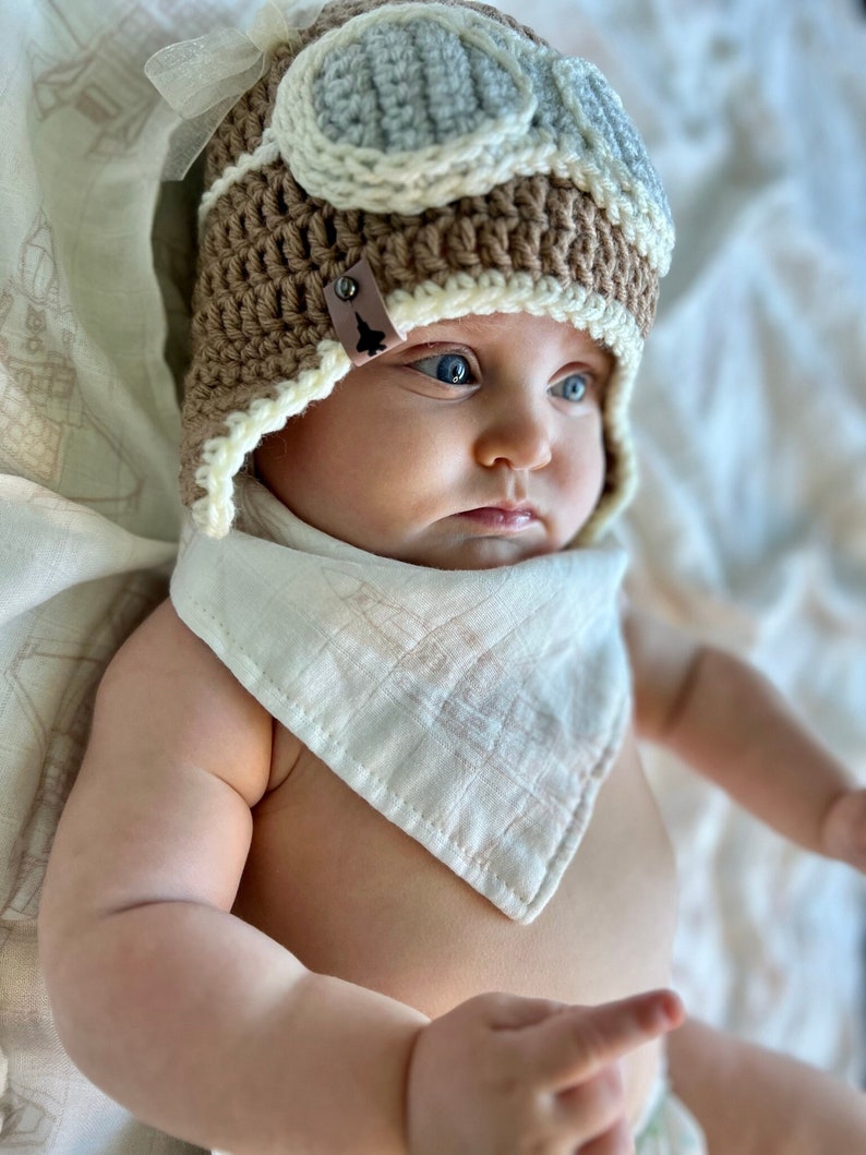 May include: A baby wearing a brown crocheted aviator hat with a white and blue faux leather goggles design. The hat has a brown leather tag with a small airplane design. The baby is wearing a white muslin bib with a subtle pink pattern.
