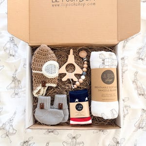 May include: An open cardboard box filled with baby items. The box contains a knitted aviator hat, a wooden airplane-shaped teether, a pacifier clip, gray booties, a pair of navy socks with white stars, and a white swaddle blanket. The box is labeled "LIL POSH Box".