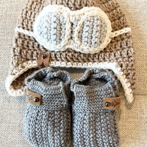 May include: A set of knitted baby booties and a crocheted hat with a faux leather aviator goggles design. The booties are gray and the hat is brown with white accents.