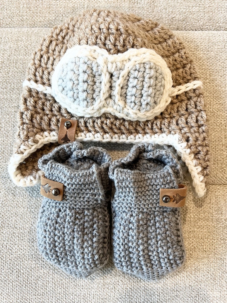 May include: A set of knitted baby booties and a crocheted hat with a faux leather aviator goggles design. The booties are gray and the hat is brown with white accents.