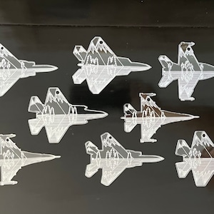 May include: Eight white acrylic cutouts of F-22 fighter jets with a mountain scene design. The cutouts are on a black surface.