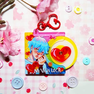 May include: A colorful charm featuring an anime-style character with blue hair and a heart design. The charm has the text "Mi Vida Loca" and is attached to a red heart-shaped clasp. Decorative buttons and pink flowers surround the charm.