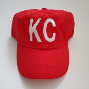 May include: A red baseball cap with white embroidered lettering that says "KC".