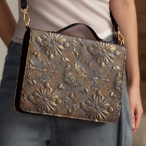 Può includere: Borsa a tracolla marrone e oro con motivo floreale in rilievo, cinturino e manico neri. La borsa ha una forma rettangolare e presenta un motivo floreale dettagliato. Il cinturino è nero con finiture color oro.