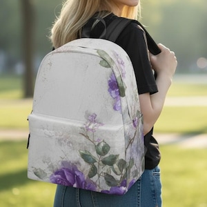 May include: A backpack featuring a light, off-white background with a watercolor-style floral print. The design includes deep purple roses, green leaves, and delicate stems scattered across the surface. It has black straps and a front pocket.