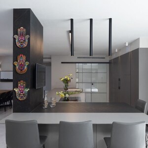 May include: Interior shot of a modern kitchen with a dining area. Three colorful Hamsa hand wall hangings are mounted on a dark gray pillar. The kitchen features a gray countertop, gray chairs, and black pendant lights.
