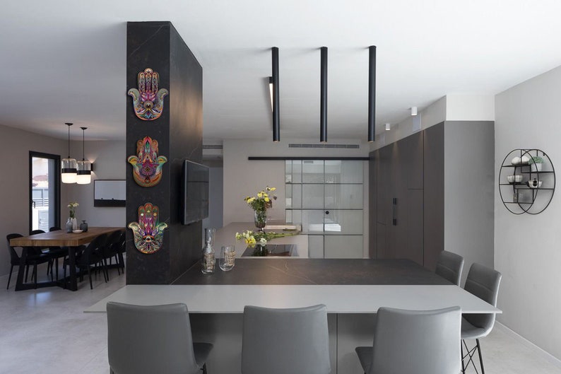 May include: Interior shot of a modern kitchen with a dining area. Three colorful Hamsa hand wall hangings are mounted on a dark gray pillar. The kitchen features a gray countertop, gray chairs, and black pendant lights.