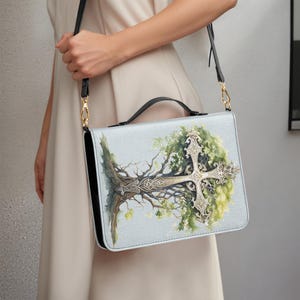 Puede incluir: Bolso bandolera azul claro con correa y asa negras. El bolso presenta una ilustración detallada de un árbol con una cruz plateada. El árbol tiene hojas verdes y ramas marrones. El bolso tiene herrajes dorados.