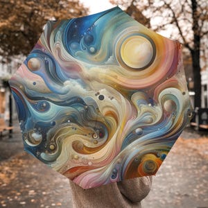 Op de afbeelding: Een abstracte kunstparaplu met een wervelend patroon in blauw, geel, oranje en roze. Het ontwerp van de paraplu bevat hemelse elementen zoals planeten en een zon, waardoor een kleurrijk en artistiek accessoire ontstaat.