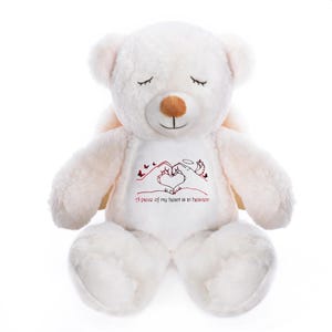 May include: A white plush teddy bear with closed eyes and a brown nose. The bear has small angel wings and a heart-shaped design made with hands on its chest, with the text "A piece of my heart is in heaven" in red.