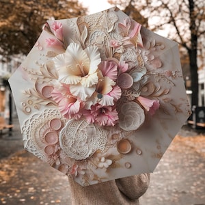 May include: An open umbrella with a floral design. The umbrella features a detailed arrangement of cream, pink, and peach-colored flowers, with intricate lace-like patterns. The background is a blurred outdoor scene with fall foliage.