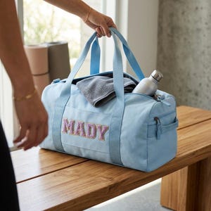 Pode incluir: Uma mala de desporto azul claro com o nome "MADY" bordado em letras rosa e roxas. Possui alças azuis claras e um bolso com zíper. Dentro, uma toalha cinza e uma garrafa de água prateada. A mala está sobre um banco de madeira.