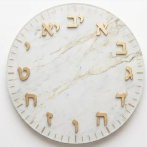 Puede incluir: Un reloj de pared redondo con una esfera de mármol blanco y números hebreos dorados. El reloj tiene un borde de vidrio transparente con pequeñas marcas de verificación. El mármol tiene sutiles vetas doradas.