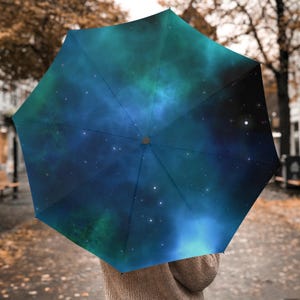 Puede incluir: Un paraguas abierto con un diseño de galaxia vibrante. La cubierta del paraguas presenta un patrón de nebulosa azul oscuro y verde azulado con estrellas blancas dispersas. El mango es de color gris oscuro.