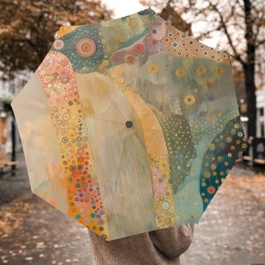 Peut inclure: Un parapluie ouvert avec un motif abstrait coloré. Le parapluie présente un motif de cercles et de formes organiques dans des tons de jaune, d'orange, de bleu et de vert. Le parapluie est tenu par une personne, avec l'arrière-plan flou.