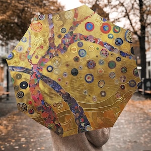 Pode incluir: Um guarda-chuva aberto com um fundo dourado e um design de árvore colorido. A árvore tem galhos com detalhes em vermelho, azul e roxo. O guarda-chuva apresenta um padrão repetitivo de círculos em várias cores.