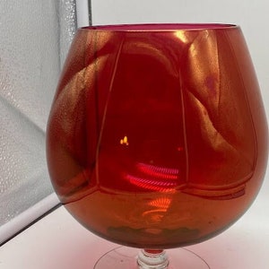 May include: A large, red glass vase with a gold iridescent finish. The vase has a round body and a wide, flared mouth. It sits on a clear glass pedestal.