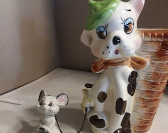 May include: A white ceramic dog figurine with black spots, wearing a green hat and a brown bow, holding a chain attached to a smaller white ceramic mouse figurine. The dog is standing next to a brown ceramic planter.