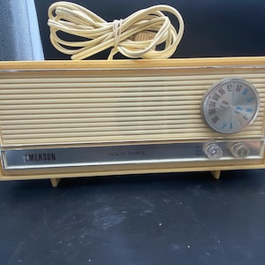 May include: A vintage cream-coloured Emerson solid state AM/FM radio with a silver face and a round dial. The radio has a cord and plug.