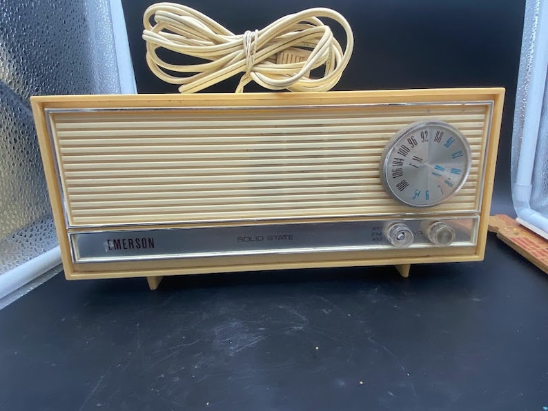 May include: A vintage cream-coloured Emerson solid state AM/FM radio with a silver face and a round dial. The radio has a cord and plug.