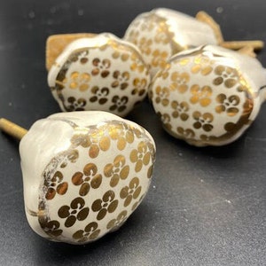 Może przedstawiać: Cztery białe ceramiczne gałki do szuflad z złotym wzorem kwiatowym. Gałki mają zaokrąglony kształt i jeden otwór na śrubę z tyłu.