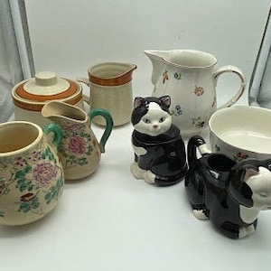 Puede incluir: Un juego de vajilla de cocina de cerámica vintage con un diseño de gato blanco y negro. El juego incluye una jarra de crema, un azucarero, una jarra y un tazón pequeño. Las piezas están decoradas con motivos florales y tienen un encanto rústico.