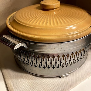 May include: A yellow ceramic casserole dish with a lid and a silver metal base with a decorative pattern. The base has a scalloped edge.