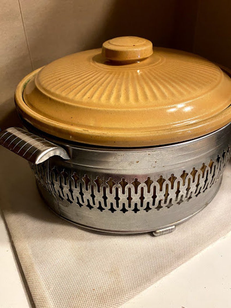 May include: A yellow ceramic casserole dish with a lid and a silver metal base with a decorative pattern. The base has a scalloped edge.