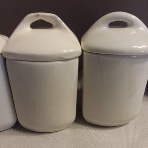 May include: Set of three white ceramic canisters with lids. Each canister has a small handle on the lid.