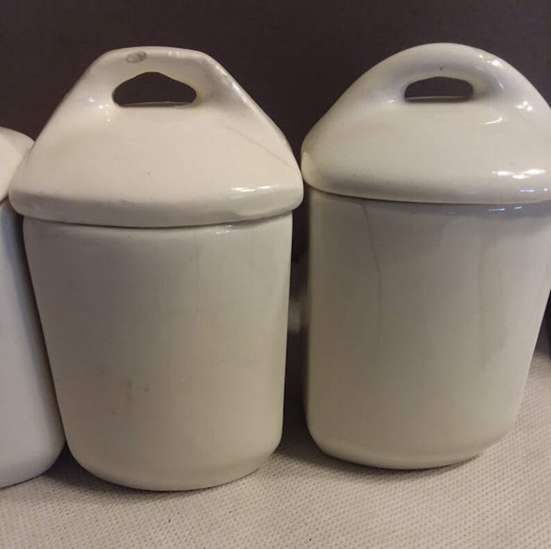 May include: Set of three white ceramic canisters with lids. Each canister has a small handle on the lid.