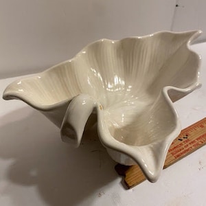May include: A white ceramic bowl shaped like a leaf with a ruffled edge. The bowl has a unique design with a curved handle.