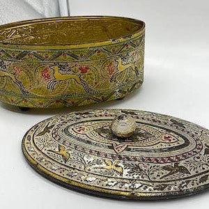 May include: An oval metal container with a removable lid, featuring a gold-toned base decorated with repeating patterns of leaping deer and foliage. The lid displays intricate floral motifs, birds, and a central spherical knob handle.