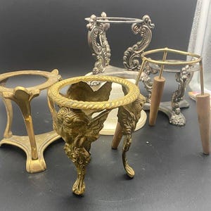May include: A collection of four decorative display stands. One is a gold-toned metal stand with three legs, another is a brass stand featuring winged figures, a third is a silver-toned ornate metal stand, and the fourth has a gold ring with wooden legs.