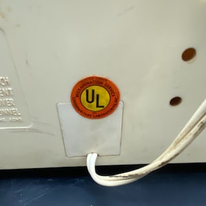 May include: A white electrical appliance with a UL certification sticker. The sticker is orange with black text and a black UL logo. The appliance has a white cord.