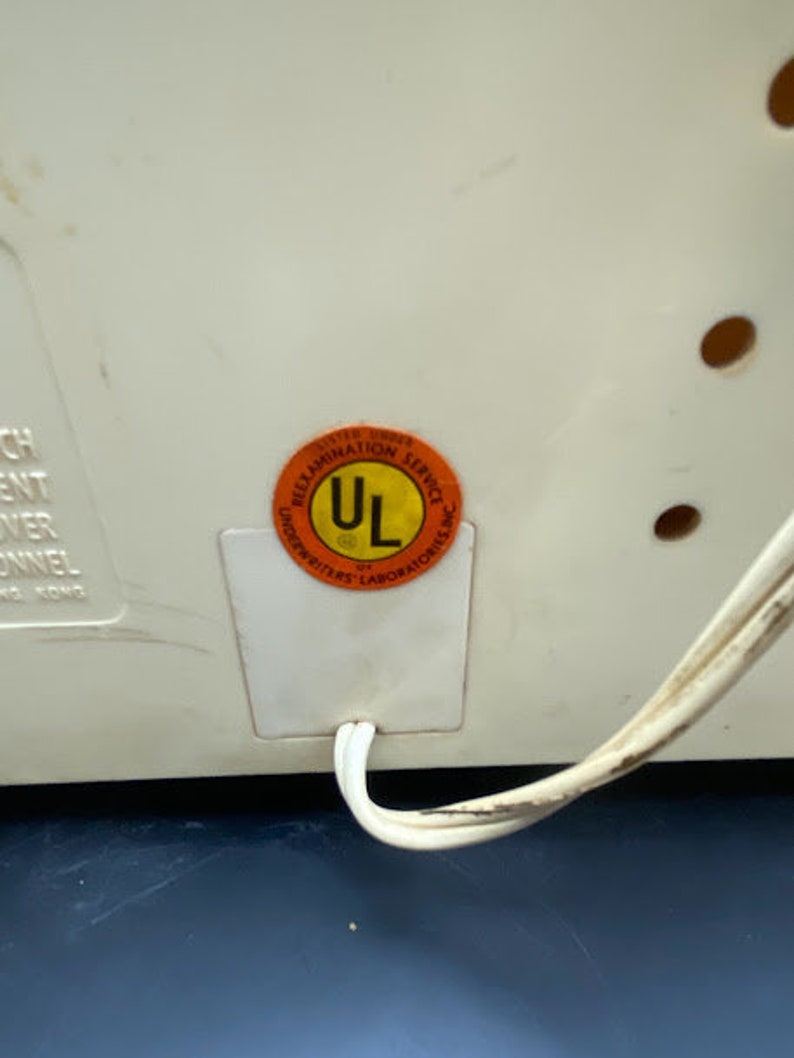 May include: A white electrical appliance with a UL certification sticker. The sticker is orange with black text and a black UL logo. The appliance has a white cord.