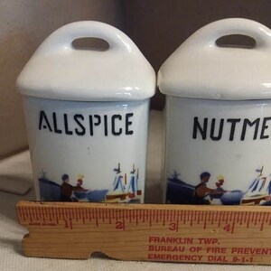 May include: Set of three white ceramic spice jars with blue and white illustrations. The jars are labelled "Allspice", "Nutmeg", and "Cinnamon".