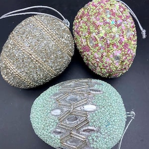May include: Three decorative egg-shaped ornaments. One is silver with a beaded design, another is covered in pink, green, and gold sequins and beads, and the third is light green with silver accents and clear gems. Each has a silver string for hanging.