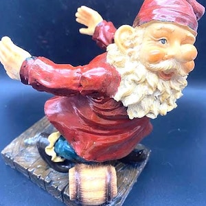 May include: A whimsical garden gnome figurine with a red hat and coat, white beard, and a smiling face. The gnome is posed with arms outstretched, standing on a wooden base with a small barrel. The figurine is painted with vibrant colors.