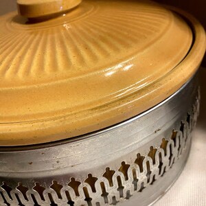 May include: A yellow ceramic casserole dish with a metal base. The base has a decorative design with a scalloped edge.