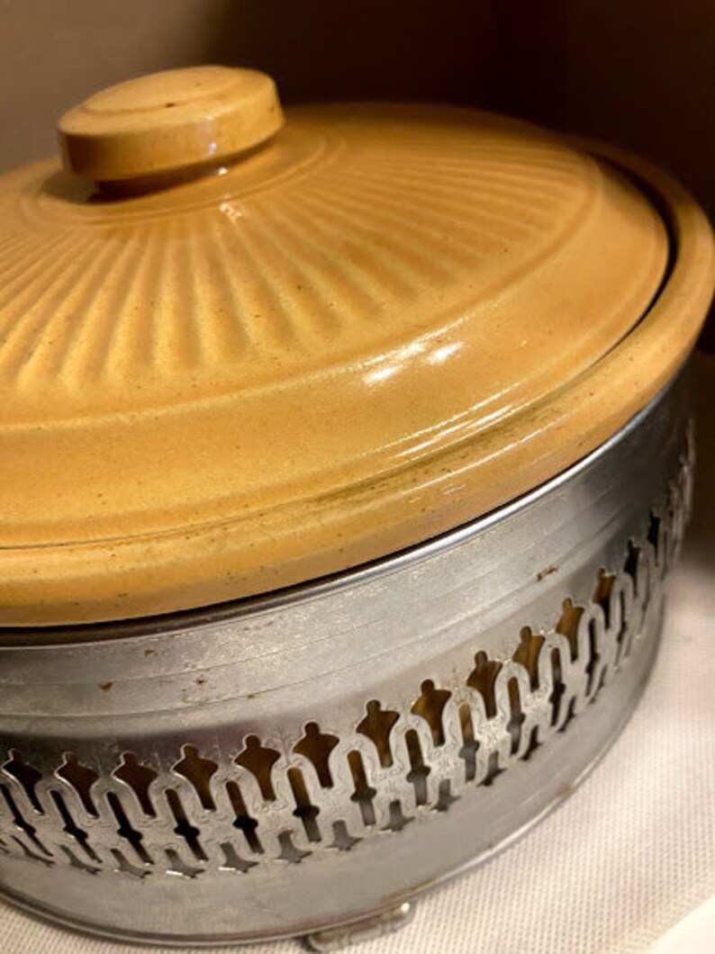 May include: A yellow ceramic casserole dish with a metal base. The base has a decorative design with a scalloped edge.