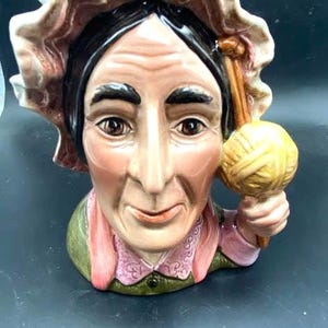May include: A ceramic figurine of an elderly woman wearing a bonnet and holding a ball of yarn. The figurine has a detailed face with rosy cheeks and brown eyes. The woman is wearing a pink scarf and a green top. The yarn is yellow.