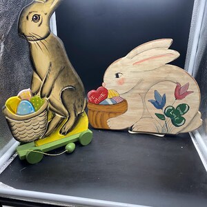 May include: Two wooden bunny figurines, one pulling a basket of colorful Easter eggs on wheels and the other holding a basket of hearts with the text "Be Mine" and a shamrock with the text "Good Luck".