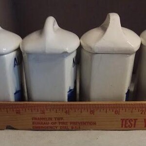 May include: Set of four white ceramic spice jars with blue and white floral designs. The jars have lids with handles and are arranged in a row.