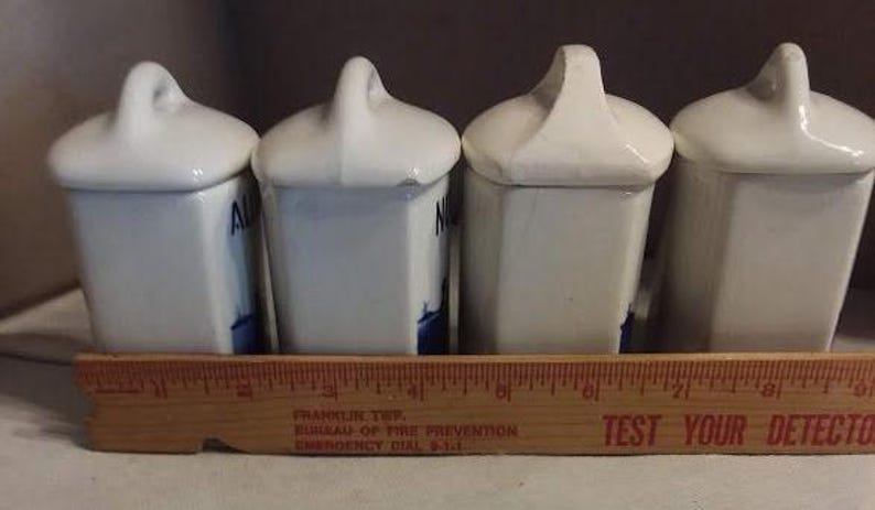 May include: Set of four white ceramic spice jars with blue and white floral designs. The jars have lids with handles and are arranged in a row.