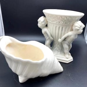 May include: Two white ceramic decorative pieces. One is a seashell-shaped planter with a light beige interior. The other is a vase with cherub figures. The vase has a detailed design and a wide opening. Both pieces are on a dark surface.